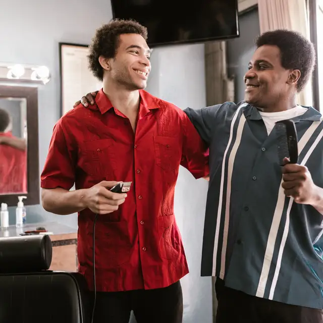 A barber with his arm around a younger barber's shoulder, mid-conversation